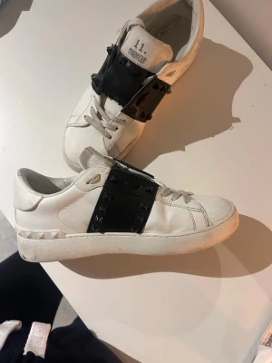 Valentino Rockstud vita sneakers - Säljer ett par vita Valentino Rockstud sneakers med svart band och nitar över vristen. Skorna har snörning, rund tå och platt sula. Tillverkade i skinn och har den ikoniska rockstud-detaljen på sidan och bak. Använda men inte trasiga någon stans.