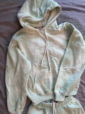 Pastelgrönt loungewear-set med hoodie - Mysigt loungewear-set i mjuk bomullsmix i en fräsch pastelgrön färg. Setet består av en hoodie med känguruficka och dragsko samt matchande byxor med resår i midjan och benslut. Perfekt för chill dagar hemma eller när du vill vara bekvämt klädd.