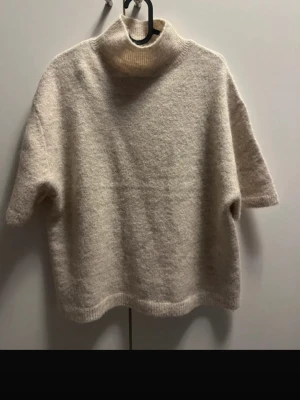 Beige stickad tröja med polokrage - Mysig beige stickad tröja med korta ärmar och hög polokrage. Tröjan har en boxy passform och är gjord i ett mjukt, fluffigt material som känns skönt mot huden. Perfekt för lager på lager och enkel att styla till olika outfits.