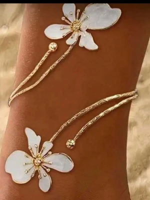 Forlänk med vita blommor - Unikt fotlänk i guldton med två stora vita blommor i emalj och guldfärgade detaljer. Den har en öppen design och tunna, böjda band som ger en elegant och trendig look. Perfekt för dig som vill sticka ut med ett smycke som känns både romantiskt och modernt.