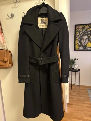 Svart trenchcoat från Burberry i ull - Säljer en svart trenchcoat från Burberry i ull med klassisk dubbelknäppning, bälte i midjan och detaljerade spännen vid ärmsluten. Kappan har bred krage och långa ärmar, perfekt för kyliga dagar. En tidlös och stilren modell med snygg passform.
