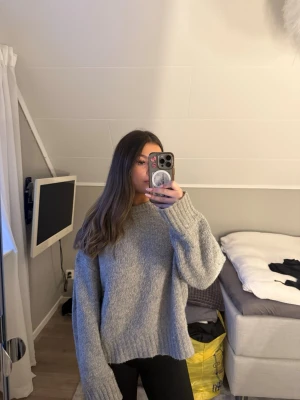 Grå oversized stickad tröja - Mysig grå stickad tröja med oversized passform och långa ärmar. Tröjan har en rund halsringning och ribbstickade muddar vid ärmslut och nederkant. 