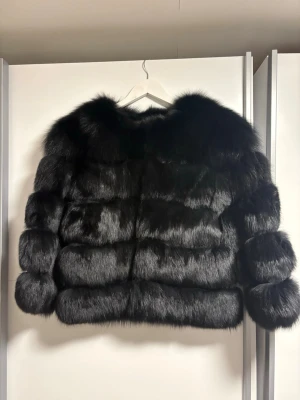 Svart fluffig pälsjacka - Snygg svart pälsjacka med fluffig och lyxig känsla. Jackan har en boxig passform och breda ärmar, perfekt för att sticka ut i mängden. Pälsen är glansig och ger en exklusiv look. Perfekt för dig som vill ha en statement-piece i garderoben.