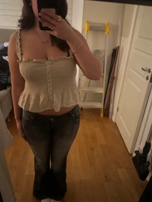 Beige volang topp med knappar - Söt beige linnetopp med volangkant och smockad midja. Toppen har breda axelband med rynkade detaljer och knappar framtill. Perfekt för varma dagar och ger en romantisk vibe. Köpt från Sellpy. Passar mig som normalt har M i toppar 