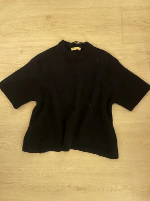 Mörkblå stickad tröja Gina Tricot - Mysig svart stickad tröja från Gina Tricot med korta ärmar och ribbad rund hals. Tröjan har en avslappnad passform och är tillverkad i ett mjukt material som känns skönt mot huden. Perfekt för lager på lager och enkel att styla till olika outfits.