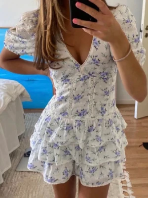 Trendig klänning - Supersöt vit klänning med blå blommönster och volanger. Puffärmar och v-ringad hals ger en romantisk vibe. Tillverkad i mjuk bomull, perfekt för varma dagar. Spetsdetaljer längs sömmarna och figurnära passform.