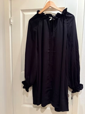 Svart blus med volangkrage från H&M - Stilren svart blus/klänning från H&M med volangkrage och knytning vid halsen. Blusen har långa ärmar med resår vid ärmslut och är tillverkad i ett lätt, flowy tyg som ger en avslappnad men chic look.