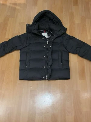 Svart Moncler dunjacka med huva - Säljer en svart Moncler dunjacka i kort modell. Jackan är varm, lätt och bekväm, perfekt för höst och vinter. Klassisk design med huva, dragkedja samt tryckknappar framtill. 
