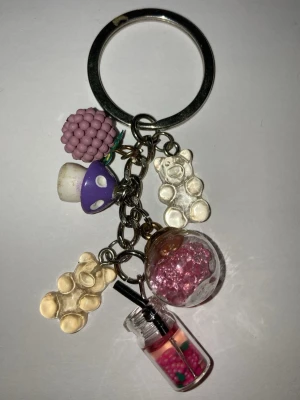 Nyckelring med söta hängen - Nyckelring som är hand gjort av mig med flera färgglada hängen: en lila hallon, två björnar i genomskinlig plast, en liten svamp, en kula med rosa pärlor och en miniatyrburk med sugrör och pärlor. Perfekt accessoar för att piffa upp väskan eller nycklarna. Frakt 22 till 100 kr beroende på vikt. Kontakta mig om du har frågor eller vill köpa.