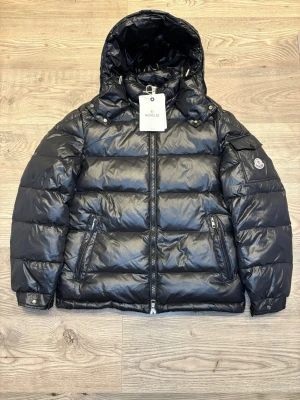 Moncler Maya - Snygg svart puffer dunjacka från Moncler med glansig finish och stor huva. Jackan har dragkedja framtill, två sidofickor med dragkedja och Moncler-logga på ärmen. Perfekt för kalla dagar och riktigt trendig look. Pris kan diskuteras