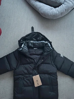 Svart burberry puffer/väst - Svart puffer dunjacka från Lindex med avtagbara ärmar så den kan användas som väst. Jackan har huva med rutigt foder, dragkedja och två framfickor med dragkedja. Perfekt för kalla dagar och riktigt snygg streetstil.