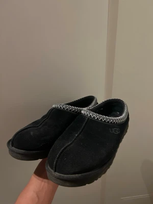 Svarta mockatofflor från UGG - Mysiga svarta tofflor från UGG i mjuk mocka med broderad kant i vitt och svart. Klassisk rund tå och platt sula, perfekta för kalla dagar. Diskret UGG-logga på sidan och skön innersula.