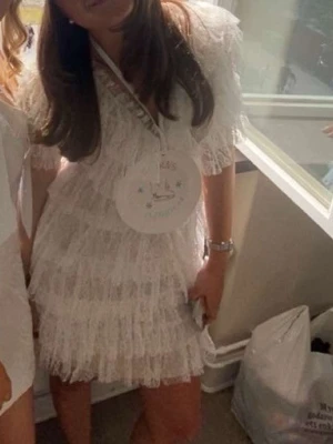 Vit spetsklänning med volanger från Malina - Drömmig kortklänning från Malina i vit spets med flera lager volanger och v-ringad hals. Klänningen har korta ärmar och en feminin siluett med romantiska detaljer. Perfekt för dig som vill sticka ut med en elegant och lekfull look.