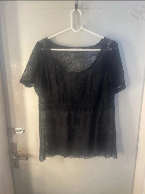 Svart spetsblus - Säljer en svart blus i genomskinlig spets med korta ärmar och rund halsringning. Blusen har dekorativa knappar framtill och ett romantiskt spetsmönster över hela plagget. Perfekt för dig som gillar en feminin och trendig stil.