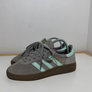 Adidas Handball Spezial grå/mint sneakers - Snygga Adidas Handball Spezial sneakers i grå mocka med mintgröna detaljer och klassiska tre ränder på sidan. Skorna har brun platt sula, snörning och guldfärgad Samba-text på sidan. Perfekta för dig som gillar retrostil och vill sticka ut.