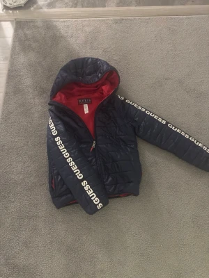 Mörkblå dunjacka från Guess - Snygg mörkblå dunjacka från Guess med huva och röd insida. Jackan har dragkedja och vita Guess-loggor längs ärmarna. Tillverkad i 100% polyester och perfekt för kyliga dagar.