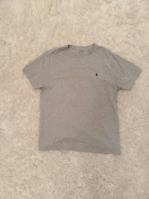 Grå t-shirt från Polo Ralph Lauren - Klassisk grå t-shirt från Polo Ralph Lauren med liten mörkblå logga broderad på bröstet. T-shirten har rund halsringning och korta ärmar. Tillverkad i mjuk bomull för en skön och avslappnad känsla. Skriv vid funderingar🙌