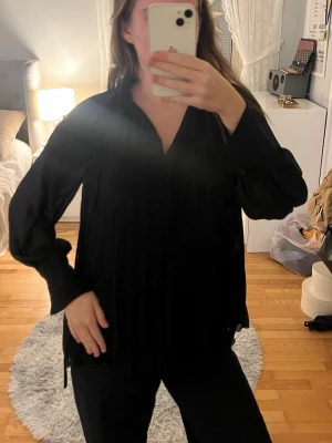 Svart plisserad blus med v-ringning - Snygg svart blus med plisserade detaljer och v-ringning. Blusen har långa ärmar med breda muddar och är tillverkad i ett lätt, något transparent tyg som ger en luftig känsla. Perfekt att styla med jeans eller kjol.