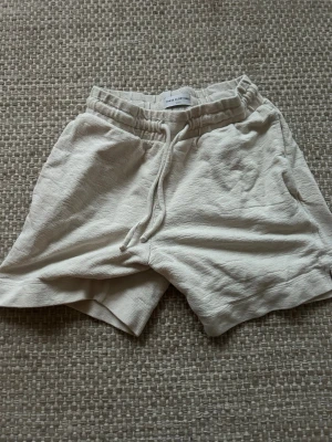 Beiga shorts från These Glory Days - Säljer ett par beiga shorts från These Glory Days med avslappnad passform. Shortsen har resårmidja med snörning och är tillverkade i ett mjukt bomullstyg. Perfekta för varma dagar och chill häng. Storlek S