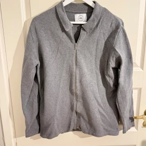 Grå ziptröja från Kronstadt Casual - Snygg grå ziptröja från Kronstadt Casual med hel dragkedja framtill och klassisk krage. Tröjan har en enkel och clean design, perfekt att slänga på sig över en t-shirt. Materialet känns mjukt och bekvämt.