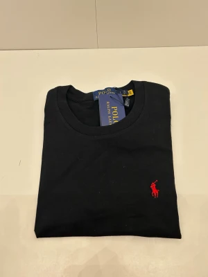 Svart t-shirt från Polo Ralph Lauren - Svart t-shirt från Polo Ralph Lauren med klassisk rund hals och ikonisk röd broderad logga på bröstet. Tillverkad i mjuk bomull som känns skön mot huden. Perfekt för en clean och stilren look. Skriv för annan info