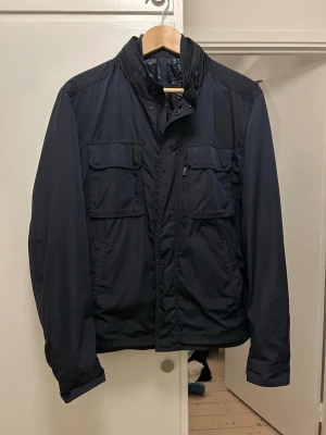 Field Jacks från Massimo dutti - Snygg mörkblå vindjacka från Massimo dutti med flera praktiska fickor framtill och hög krage. Jackan har dragkedja och tryckknappar, samt långärmade ärmar. Perfekt för blåsiga dagar och enkel att matcha med olika outfits.