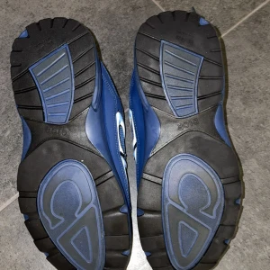 Blå Dior B30 sneakers med snörning - Säljer ett par blå Dior B30 sneakers med coola detaljer och snörning. Skorna har mesh och syntetpaneler, samt reflekterande vita och svarta linjer längs sidorna. CD-loggan syns tydligt på sidan och sulan är robust med mönster för extra grepp.