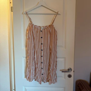 Randig sommarklänning med knappar - Säljer en vit och beige randig klänning med smala axelband och knappar framtill. Klänningen har en avslappnad passform och är perfekt för varma dagar. Materialet känns lätt och luftigt, vilket gör den skön att bära.