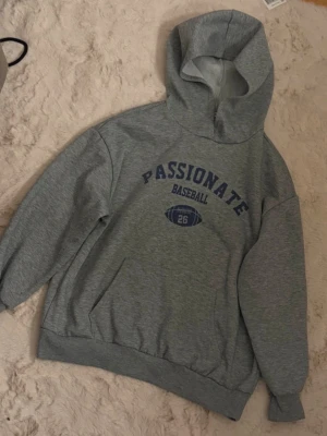Grå hoodie från SHEIN Teen - Grå hoodie från SHEIN Teen med blå text 'PASSIONATE BASEBALL' och en amerikansk fotboll på bröstet. Klassisk känguruficka framtill och mjuk insida. Perfekt för chill dagar och enkel att matcha med jeans eller mjukisbyxor.