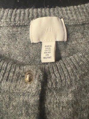 Grå kofta från H&M med guldknappar - Mysig grå kofta från H&M i storlek XL. Koftan har rund halsringning, ribbade muddar och stängs med snygga guldknappar framtill. Perfekt att slänga över en t-shirt eller topp för en chill och stilren look.