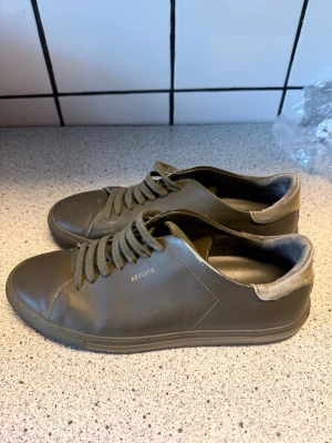Gröna sneakers från Axel Arigato - Snygga gröna sneakers från Axel Arigato med minimalistisk design och guldfärgad logga på sidan. Skorna har snörning, rund tå och är tillverkade i skinn med mjuk vadderad kant i grått. Perfekta för en clean och stilren look.