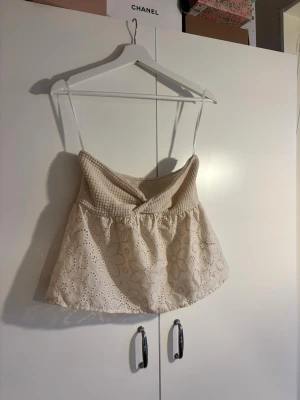 Beige broderad linnetopp med spets - Söt beige linnetopp med tunna axelband och broderade blommor. Överdelen är smockad och ger en skön passform, medan nederdelen har volang och spetsdetaljer. 