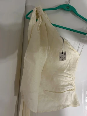 Krämvit oneshoulder topp Zara  - Säljer en krämvit oneshoulder topp i linne med lång knytband på axeln och dekorativa fransar längst ut. Toppen har lätt rynkning i sidan för en snygg passform och är perfekt för sommarens varma dagar.