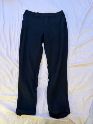 Svarta kostymbyxor med resårmidja - Säljer ett par svarta kostymbyxor med rak passform och resår i midjan. Byxorna har uppvikta benslut och bälteshällor. Perfekta till en clean och stilren look. Materialet känns mjukt och bekvämt, troligtvis polyester eller bomull.