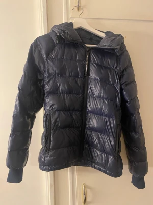 Mörkblå dunjacka Peak Performance - Snygg mörkblå dunjacka från Peak Performance med huva och dragkedja. Jackan har quiltad design, långa ärmar med muddar och två fickor med dragkedja. Perfekt för kalla dagar och enkel att matcha med olika outfits.