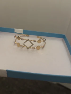 Guldigt armband med cirklar och stenar - Unikt guld platerat armband med dubbla band, dekorerat med fyra blanka cirklar och en fyrkant i mitten täckt av små gnistrande stenar. Armbandet har en öppen design och detaljer i metall som ger en modern och trendig look.