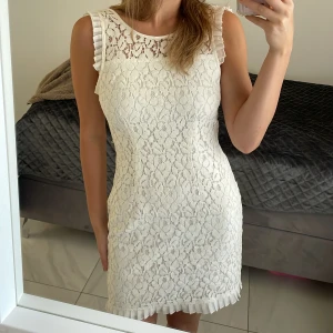 Klänning - Vit klänning i spets med blommigt mönster och volangdetaljer vid axlar och nederkant.