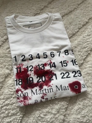 Vit t-shirt Maison Martin Margiela - Vit t-shirt från Maison Martin Margiela med svart kalendertryck och röda färgdetaljer framtill. Klassisk rund halsringning och korta ärmar. Tillverkad i 100% bomull för en skön och luftig känsla. Vill få såld snabbt passar runt 180cm eller kortare