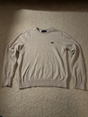 Beige stickad tröja från Lexington - Snygg beige stickad tröja från Lexington med rund halsringning och diskret logga på bröstet. Tröjan har ribbade muddar vid ärmslut och nederkant, vilket ger en klassisk och clean look. Perfekt för dig som gillar stilrena plagg i mjukt material. Skriv vid funderingar!