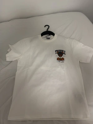 Vit t-shirt med björntryck - Vit t-shirt  med stort tryck av en björn i rånarluva och texten 'TROUBLE' på ryggen. Framsidan har ett mindre tryck på bröstet. Skön passform och cool streetstil, perfekt för dig som gillar unika motiv.