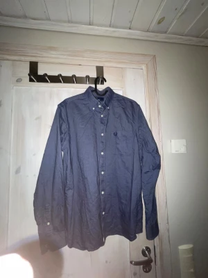 Mörkblå skjorta från Fred Perry - Snygg mörkblå skjorta från Fred Perry med klassisk button-down krage och vita knappar. Skjortan har lång ärm och broderad logga på bröstet. Perfekt för en stilren och avslappnad look. Priset går att diskuteras.