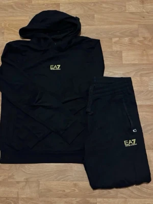 Svart EA7 set med hoodie och byxor - Snyggt svart set från EA7 Emporio Armani med hoodie och matchande byxor. Båda plaggen har EA7-logga i gult framtill. Hoodien har huva och byxorna har dragsko i midjan samt fickor. Perfekt för en avslappnad och sportig stil.