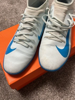 Nike innomhusskor academy futsal - Snygga fotbollsskor från Nike i vitt med blå detaljer och blå sula. Skorna har snörning och är tillverkade i syntetmaterial med en modern, sportig design. Perfekta för dig som vill sticka ut på planen med stilrena och funktionella skor.