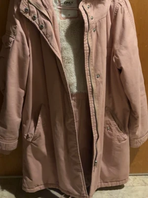 Rosa parkas från ONLY med fuskpälsfoder - Mysig rosa parkajacka från ONLY med fluffigt fuskpälsfoder på insidan. Jackan har huva, dragkedja och knappar framtill samt stora fickor och detaljer på ärmen. Perfekt för kyliga dagar och ger en chill vibe. VIKTIGT!! Den är L i storlek så den är mindre än va de står den är
