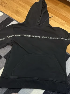 Svart hoodie från Calvin Klein Jeans - Snygg svart hoodie från Calvin Klein Jeans med logga tryckt i vitt längs bröstet och armarna. Klassisk känguruficka framtill och justerbar huva med dragsko. Tillverkad i mjukt bomullsmaterial som känns skönt mot huden. Perfekt för en avslappnad och trendig look.