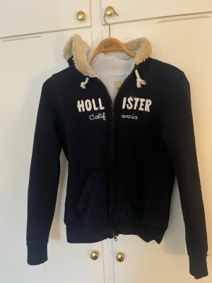Marinblå hoodie från Hollister - Mysig marinblå hoodie från Hollister med dragkedja och vit broderad text på bröstet. Fodrad med mjuk, beige teddy på insidan av huvan. Två fickor framtill och ribbade muddar vid ärmar och nederkant. Perfekt för chill dagar.
