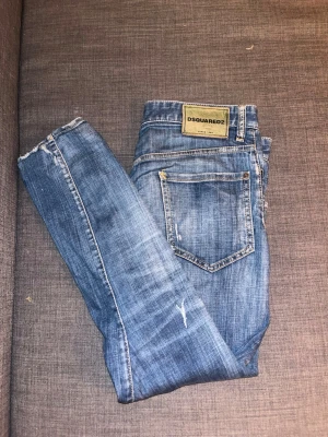 Blå Dsquared2 jeans, storlek 48 - Säljer ett par blå Dsquared2 jeans i storlek 48. Jeansen har en klassisk femficksmodell, snygga slitningar och en ljus tvätt. Tillverkade i Italien av bomullsmaterial. Perfekta för dig som gillar trendiga och bekväma jeans med en modern passform.