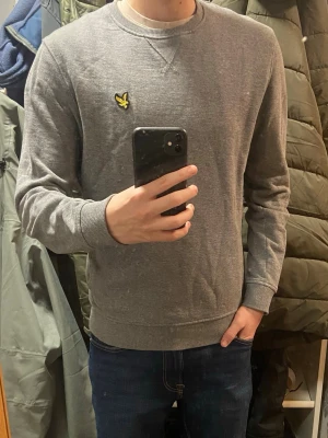 Grå sweatshirt från Lyle & Scott - Säljer en stilren grå sweatshirt från Lyle & Scott i ekologisk bomull. Tröjan har rund hals, ribbade muddar och den klassiska gula loggan på bröstet. Perfekt för en chill och avslappnad look.