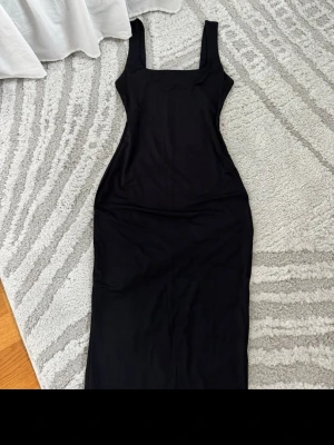 Svart långklänning med breda axelband - En stilren svart långklänning med breda axelband och rak skärning. Klänningen har en enkel, elegant design och är figurnära. Perfekt för dig som gillar minimalistisk stil. Materialet känns mjukt och stretchigt, troligen bomull eller polyester.