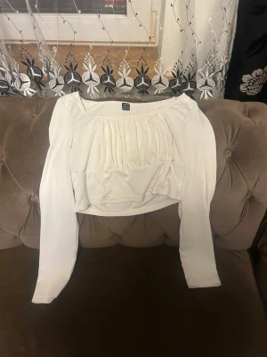 Vit croppad långärmad topp - Trendig vit croppad topp med långa ärmar och rynkade detaljer framtill. Toppen har en fyrkantig ringning och är tillverkad i ett ribbat material som ger en snygg och bekväm passform. Perfekt att styla med jeans eller kjol.